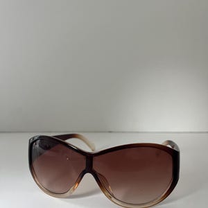Pu&ograve; includere: Occhiali da sole marroni oversize con lenti sfumate. Gli occhiali hanno una montatura marrone scuro che sfuma in un marrone pi&ugrave; chiaro nella parte inferiore. Le aste sono di colore marrone chiaro.