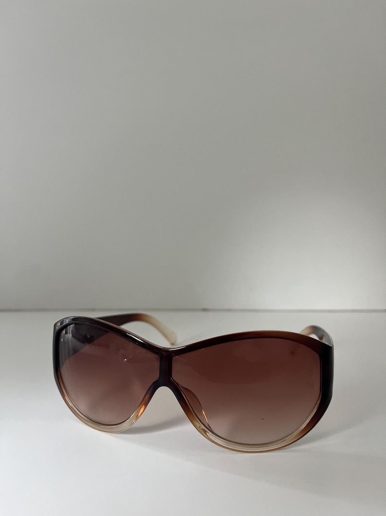 Pu&ograve; includere: Occhiali da sole marroni oversize con lenti sfumate. Gli occhiali hanno una montatura marrone scuro che sfuma in un marrone pi&ugrave; chiaro nella parte inferiore. Le aste sono di colore marrone chiaro.