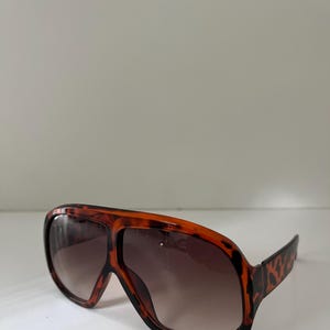 May include: Oversized sunglasses with a brown and black tortoiseshell pattern. The lenses are gradient, transitioning from dark brown at the top to a lighter shade at the bottom. The frames are thick and angular, with a retro aesthetic.