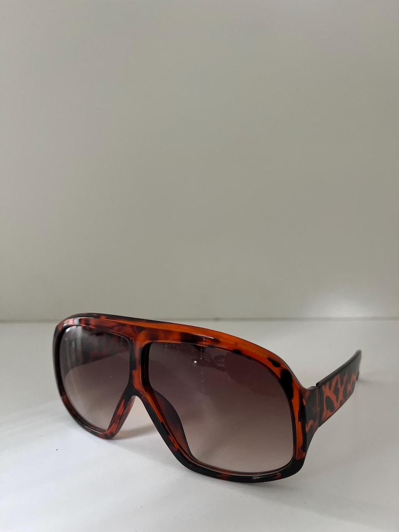 May include: Oversized sunglasses with a brown and black tortoiseshell pattern. The lenses are gradient, transitioning from dark brown at the top to a lighter shade at the bottom. The frames are thick and angular, with a retro aesthetic.