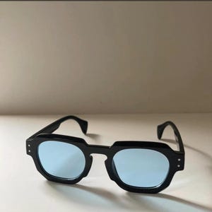 May include: Black sunglasses with a square frame and light blue lenses. The sunglasses have a thick, angular frame and a classic design. The lenses are a light blue color, and the frame is black.