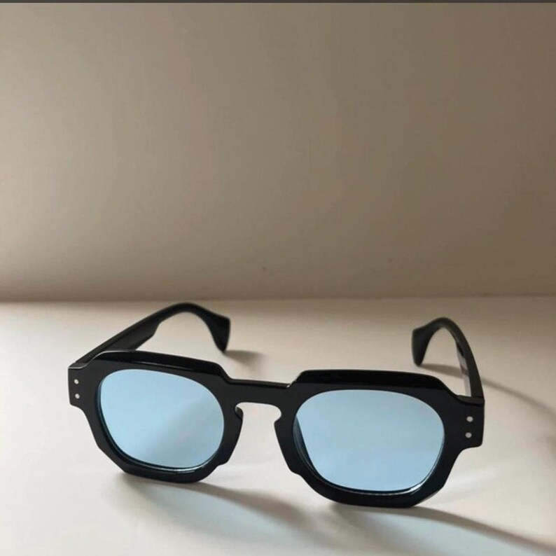 May include: Black sunglasses with a square frame and light blue lenses. The sunglasses have a thick, angular frame and a classic design. The lenses are a light blue color, and the frame is black.
