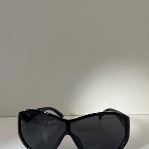 Pu&ograve; includere: Occhiali da sole neri con una montatura ampia e curva e lenti scure. Gli occhiali da sole hanno un design elegante e moderno. La montatura &egrave; spessa e sembra essere realizzata in un materiale lucido.