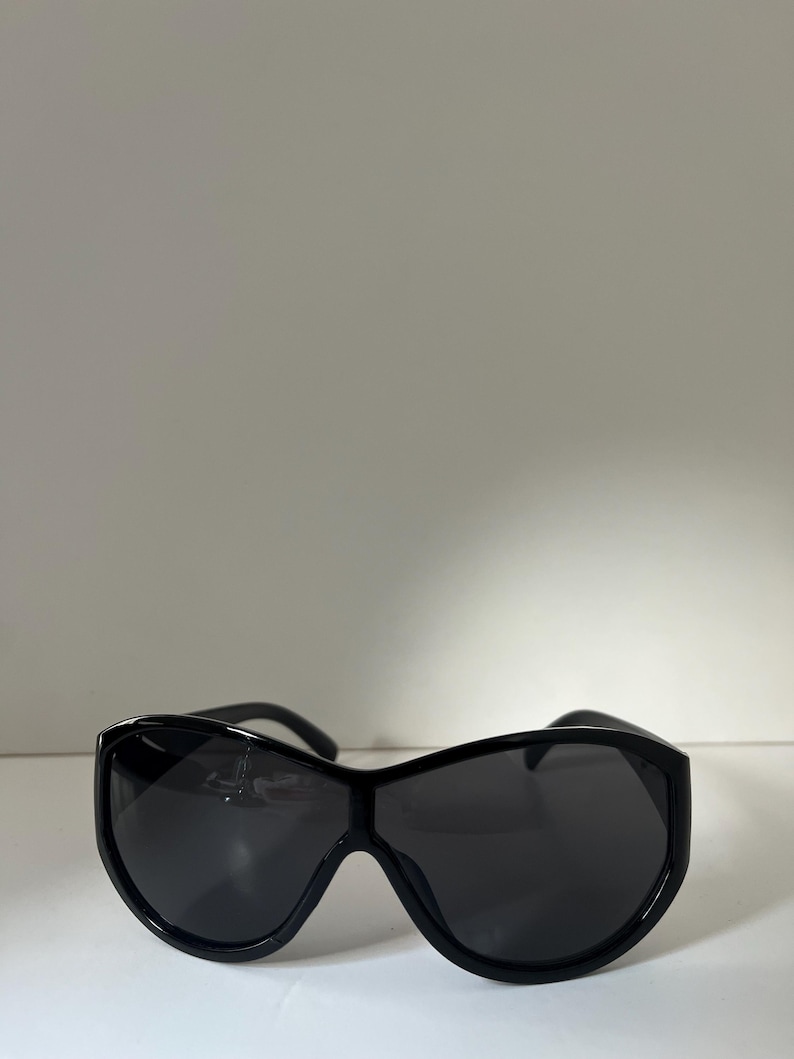 Pu&ograve; includere: Occhiali da sole neri con una montatura ampia e curva e lenti scure. Gli occhiali da sole hanno un design elegante e moderno. La montatura &egrave; spessa e sembra essere realizzata in un materiale lucido.