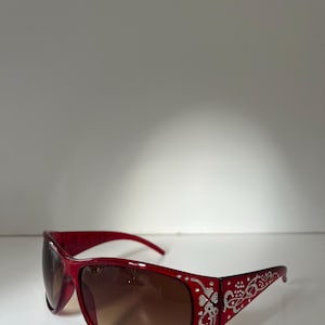 May include: Red sunglasses with a decorative silver floral design on the temples and small clear rhinestones. The lenses have a gradient tint, transitioning from brown at the top to a lighter shade at the bottom. The frames are a glossy red.