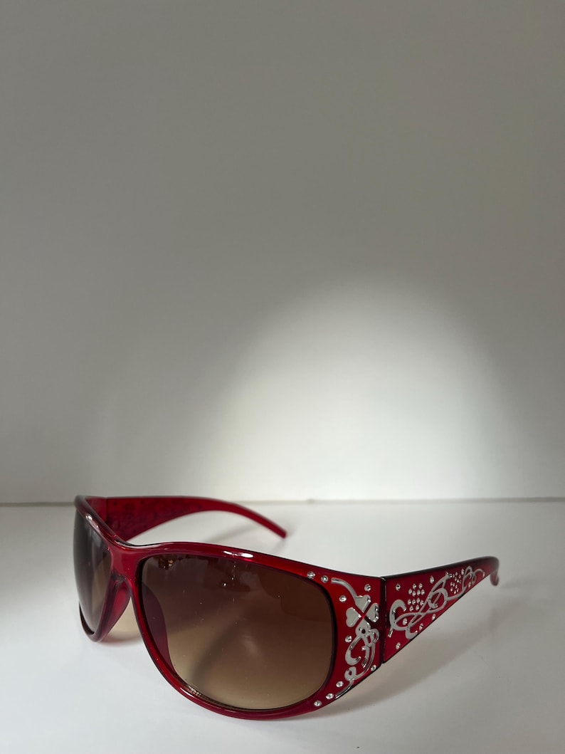 May include: Red sunglasses with a decorative silver floral design on the temples and small clear rhinestones. The lenses have a gradient tint, transitioning from brown at the top to a lighter shade at the bottom. The frames are a glossy red.