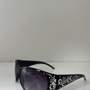 May include: Black sunglasses with a decorative silver design on the temples. The lenses are a gradient from dark purple to clear. The sunglasses have a curved shape and are set against a white background.