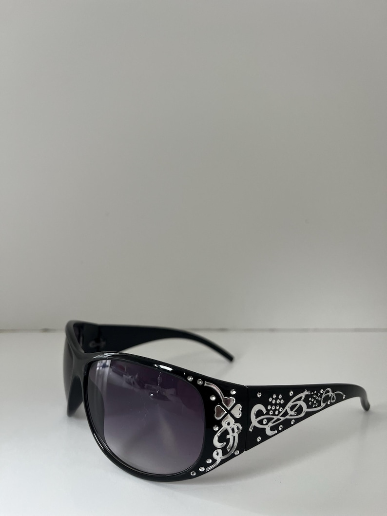 May include: Black sunglasses with a decorative silver design on the temples. The lenses are a gradient from dark purple to clear. The sunglasses have a curved shape and are set against a white background.