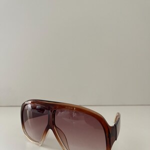 May include: Brown aviator-style sunglasses with a gradient lens. The frames are a translucent brown color, and the lenses transition from a light brown at the bottom to a darker brown at the top. The sunglasses have a classic, oversized design.