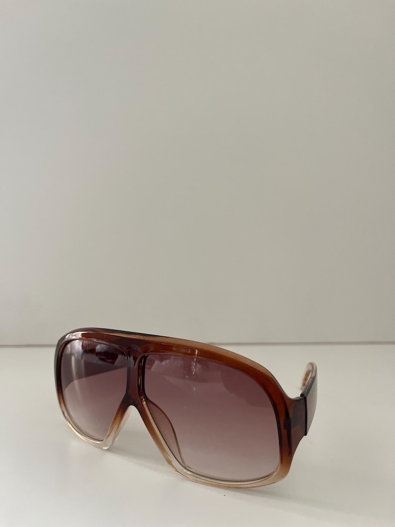 May include: Brown aviator-style sunglasses with a gradient lens. The frames are a translucent brown color, and the lenses transition from a light brown at the bottom to a darker brown at the top. The sunglasses have a classic, oversized design.