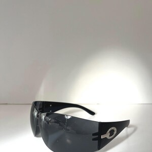 May include: Black sunglasses with a wraparound design. The sunglasses have a silver accent with a circular design on the temples. The lenses are dark, and the frames are black.