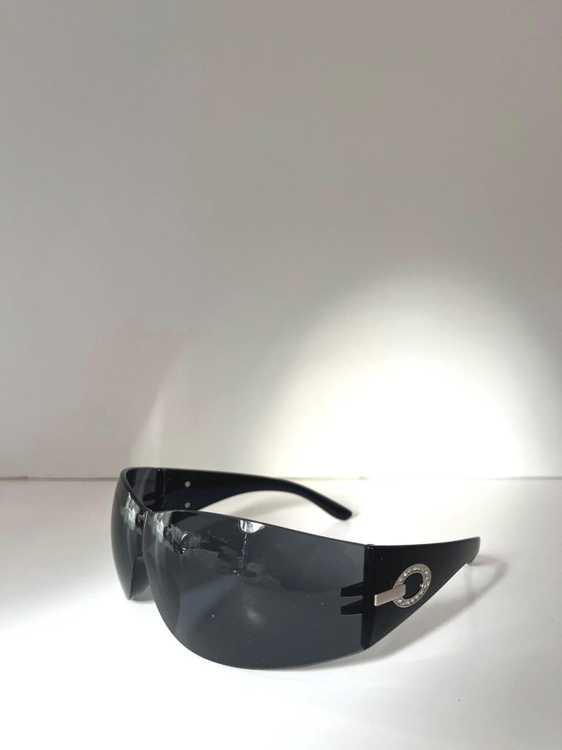 May include: Black sunglasses with a wraparound design. The sunglasses have a silver accent with a circular design on the temples. The lenses are dark, and the frames are black.