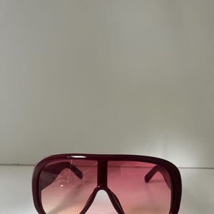 May include: Oversized burgundy sunglasses with a gradient pink and orange tint. The sunglasses have a thick frame and a bold, retro design, offering a statement look. The lenses transition from a light orange to a vibrant pink.