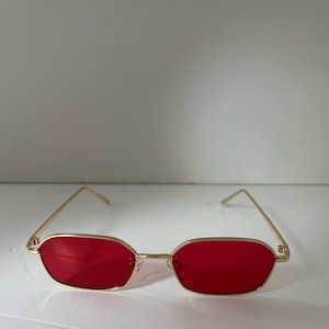 May include: Gold-framed sunglasses with rectangular red lenses. The glasses have a thin gold frame and a classic design. The lenses are a vibrant red colour, offering a bold contrast to the gold frame. These sunglasses are a stylish accessory.