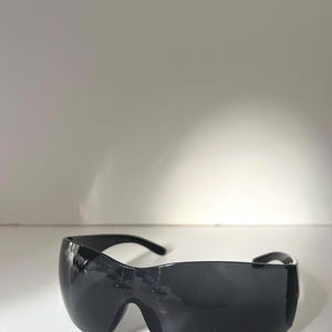 May include: Black wraparound sunglasses with a curved, shield-like design. The lenses are dark, and the frame is a matching black. The sunglasses have a sleek, modern aesthetic.