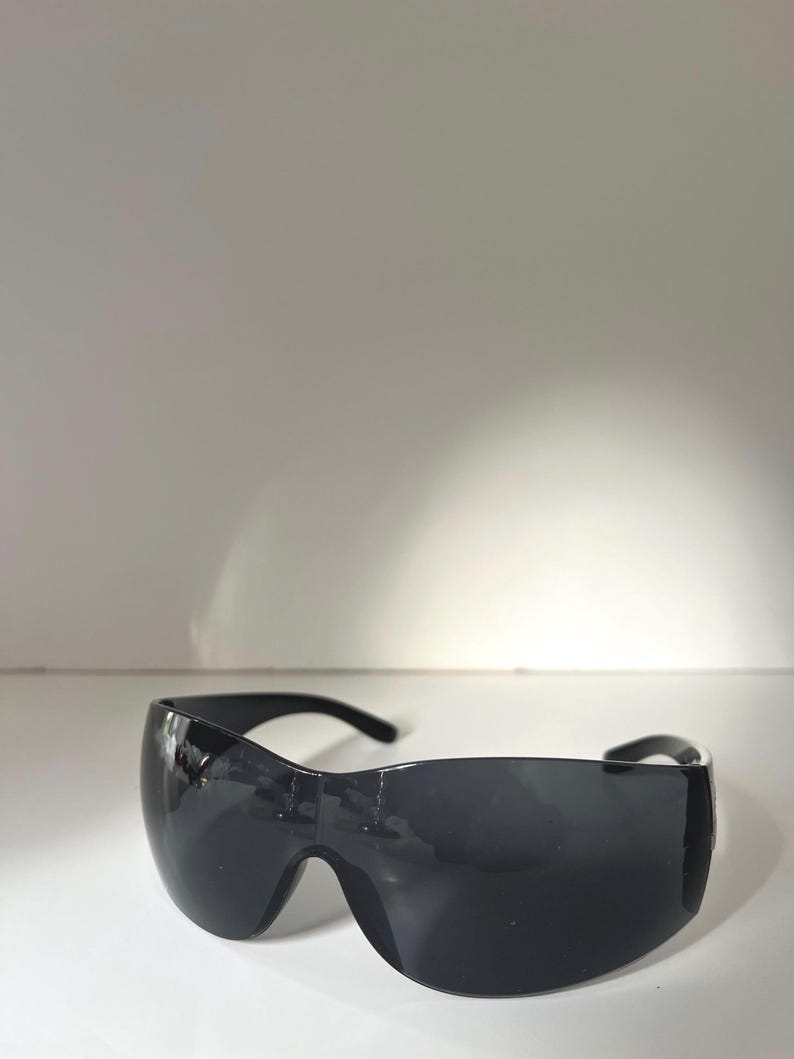 May include: Black wraparound sunglasses with a curved, shield-like design. The lenses are dark, and the frame is a matching black. The sunglasses have a sleek, modern aesthetic.
