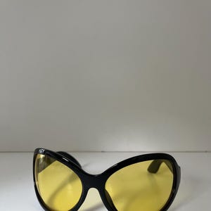 May include: Black sunglasses with large, yellow-tinted lenses. The sunglasses have a rounded, oversized shape, offering a bold and retro aesthetic. The lenses are a vibrant yellow, contrasting with the black frame.