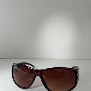 May include: Brown sunglasses with a dark frame and gradient lenses. The sunglasses have decorative embellishments on the temples. The lenses are a gradient brown color.