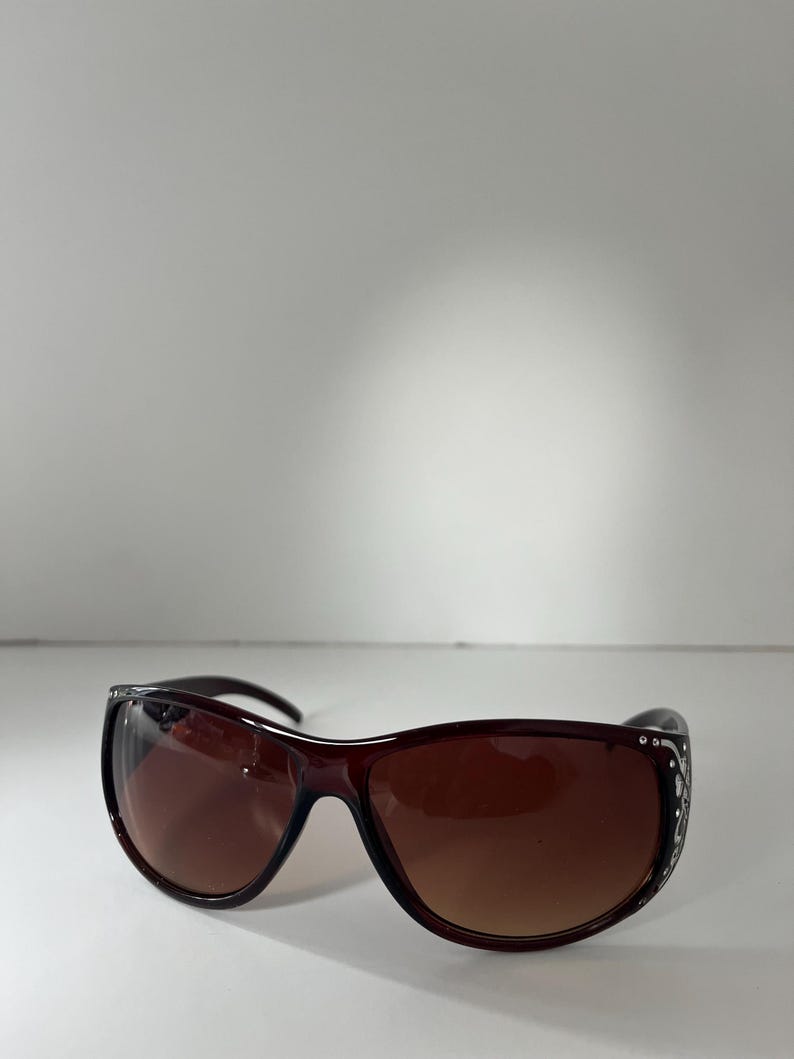May include: Brown sunglasses with a dark frame and gradient lenses. The sunglasses have decorative embellishments on the temples. The lenses are a gradient brown color.