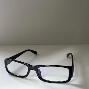 May include: Black rectangular spectacles with clear lenses. The glasses have a thick, glossy black frame and curved arms. The lenses appear to have a blue tint. The spectacles are set against a white background.
