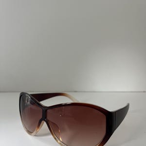 Pu&ograve; includere: Occhiali da sole marroni oversize con lenti sfumate. Gli occhiali hanno una montatura marrone scuro che sfuma in un marrone pi&ugrave; chiaro nella parte inferiore delle lenti. Le aste sono marrone scuro.