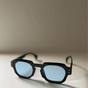 May include: Black sunglasses with a geometric frame and light blue lenses. The sunglasses have a modern design with a thick, angular frame and a subtle, minimalist aesthetic. The lenses provide a cool, stylish look.