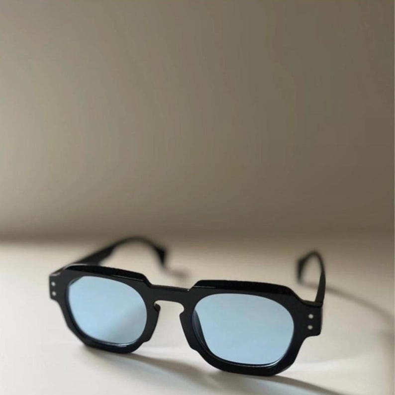 May include: Black sunglasses with a geometric frame and light blue lenses. The sunglasses have a modern design with a thick, angular frame and a subtle, minimalist aesthetic. The lenses provide a cool, stylish look.