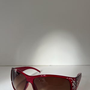 May include: Red sunglasses with brown gradient lenses. The frames are a vibrant red with decorative silver accents along the temples. The sunglasses have a sleek, modern design, suitable for sunny days.