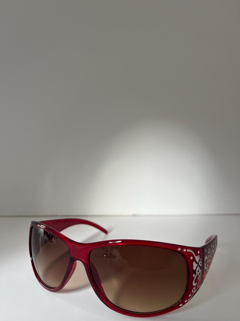 May include: Red sunglasses with brown gradient lenses. The frames are a vibrant red with decorative silver accents along the temples. The sunglasses have a sleek, modern design, suitable for sunny days.