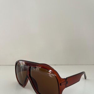 May include: Brown aviator sunglasses with a rectangular frame and dark lenses. The sunglasses have a classic design with a thick, transparent frame and straight temples. The lenses are a dark brown color.