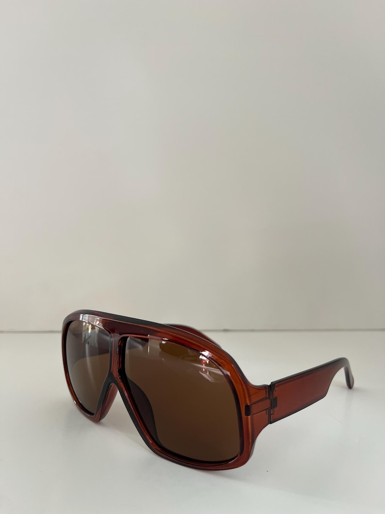 May include: Brown aviator sunglasses with a rectangular frame and dark lenses. The sunglasses have a classic design with a thick, transparent frame and straight temples. The lenses are a dark brown color.