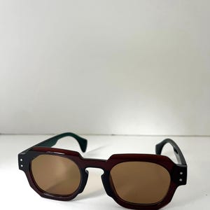 May include: Brown sunglasses with a geometric frame and brown lenses. The temples are black with green tips. The sunglasses have a modern, angular design.