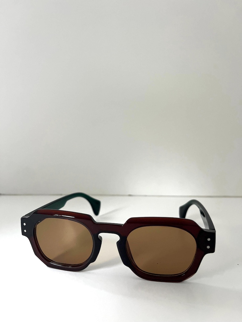 May include: Brown sunglasses with a geometric frame and brown lenses. The temples are black with green tips. The sunglasses have a modern, angular design.