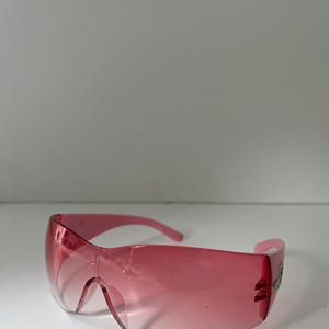 May include: Pink wraparound sunglasses with a gradient lens. The sunglasses have a curved design and a decorative element on the temple. The frame and lenses are a matching shade of pink.