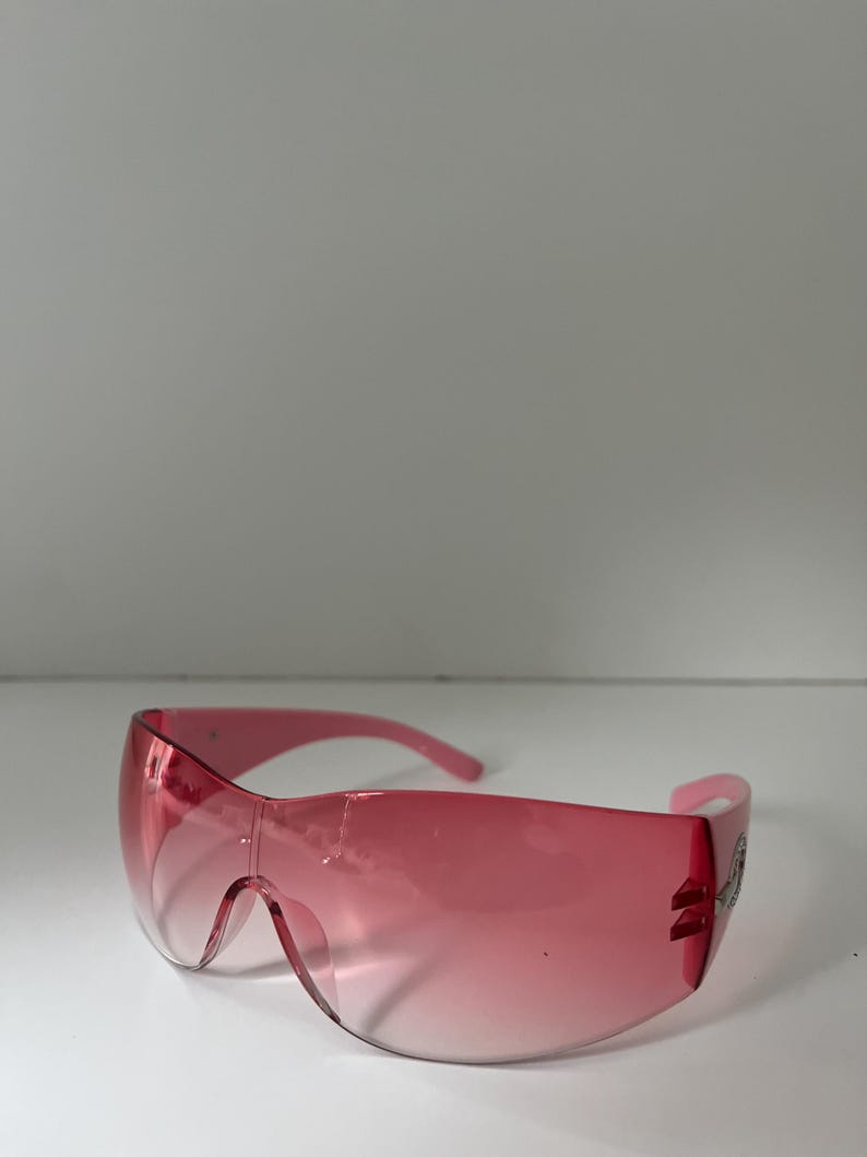 May include: Pink wraparound sunglasses with a gradient lens. The sunglasses have a curved design and a decorative element on the temple. The frame and lenses are a matching shade of pink.