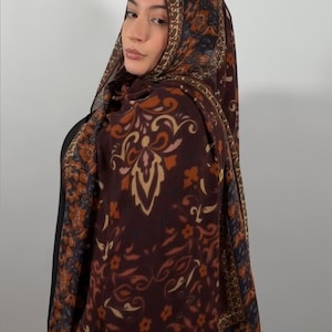 May include: A rectangular scarf featuring a dark brown base with a repeating floral and paisley pattern in shades of orange, tan, and muted blue. The scarf has a wide decorative border with a dense floral print and a thin, dark blue edge trim.
