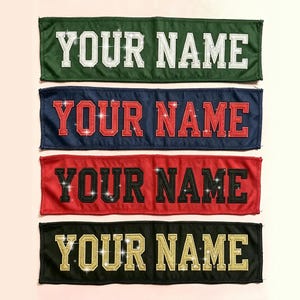 May include: Four rectangular fabric patches featuring the text YOUR NAME in block lettering. The top patch is dark green with white text, the second is navy blue with red text, the third is red with black text, and the bottom is black with gold text.