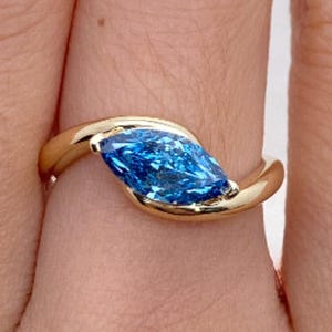 May include: A gold ring featuring a marquise-cut blue gemstone set in a bypass style band. The metal band curves around the central stone, creating a sleek and continuous design that highlights the vibrant blue color of the faceted marquise gem.