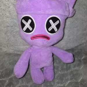 May include: A plush, lavender-colored doll with a stylized face. The doll has black eyes with white 'X' markings, a pink mouth, and a small, hat-like head covering. The doll has arms and legs and is made of a soft, fuzzy material.