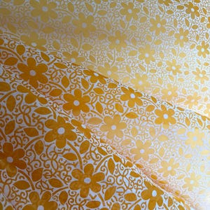 May include: A sheet of yellow and white patterned paper featuring a repeating floral design. The surface displays a dense arrangement of five-petaled flowers intertwined with leafy vines and delicate scrollwork, creating a continuous botanical motif.