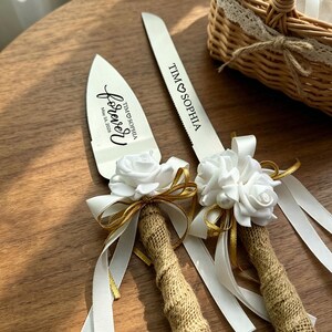 May include: A silver cake knife and server set featuring burlap-wrapped handles, white fabric roses, gold ribbon accents, and white satin ribbons. The blades are engraved with the text TIM HEART SOPHIA and the word forever with May 16, 2026.