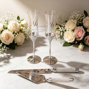 May include: Two clear glass champagne flutes with silver stems featuring crystal-encrusted bases, paired with a matching silver cake knife and server set. The flutes are engraved with the text Mr and Mrs, while the cake server features the text Ashley and Kim.