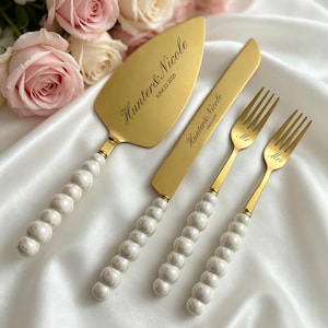 Peut inclure: Un ensemble doré de serveur à gâteau, couteau et deux fourchettes pour mariage. Le serveur et le couteau sont gravés de noms et d'une date. Les fourchettes sont gravées de "Mr" et "Mrs". Les manches sont blancs avec un motif perlé.