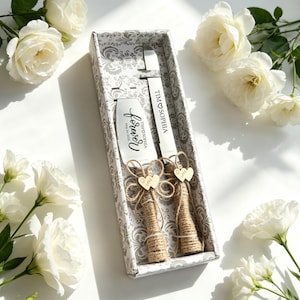 May include: A wedding cake serving set in a decorative box. The set includes a cake server and knife, both with the names "TIM & SOPHIA" and the word "forever". The handles are wrapped in twine and adorned with wooden hearts. White roses surround the box.