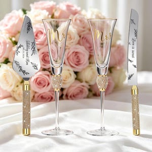 Op de afbeelding: Een taartschep, twee champagneglazen en een taartmes. De schep en het mes hebben gouden handvatten ingelegd met heldere kristallen en zilveren bladen met zwarte tekst Ashley and Kim 09.23.2026 en Love is sweet. De glazen hebben kristallen stelen.