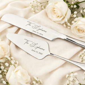 Könnte beinhalten: Ein silbernes Kuchenmesser- und Servier-Set. Das Messer hat eine gerade Klinge, und der Servierer hat eine gezahnte Kante. Beide sind mit "The Lykasons" und "June 21, 2026" graviert. Weiße Rosen und Schleierkraut umgeben das Set.
