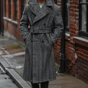 May include: A long, double-breasted coat in a grey and black herringbone pattern. The coat features a wide collar, a matching fabric belt with a rectangular buckle, and two rows of dark buttons. It is worn over dark trousers and black boots.
