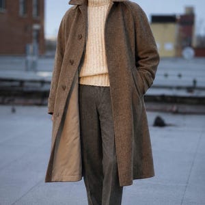 Könnte beinhalten: Ein brauner Wollmantel mit Fischgrätenmuster und breitem Kragen, ein cremefarbener Rollkragenpullover aus Rippstrick, eine dunkelgraue Wollhose mit Umschlag, braune Lederschuhe und eine passende braune Schiebermütze mit Fischgrätenmuster.
