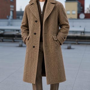 Puede incluir: Un abrigo largo de color tostado con estampado de espiga, solapa ancha, tres botones oscuros y dos bolsillos con solapa. El abrigo se lleva sobre un suéter marrón oscuro con cremallera, camisa blanca, pantalones tostados y zapatos de cuero.