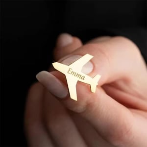Puede incluir: Un pin dorado en forma de avión con el nombre "Emma" grabado en el cuerpo. El pin se sostiene entre los dedos sobre un fondo oscuro. El pin tiene alas y cola, pareciendo un avión pequeño.